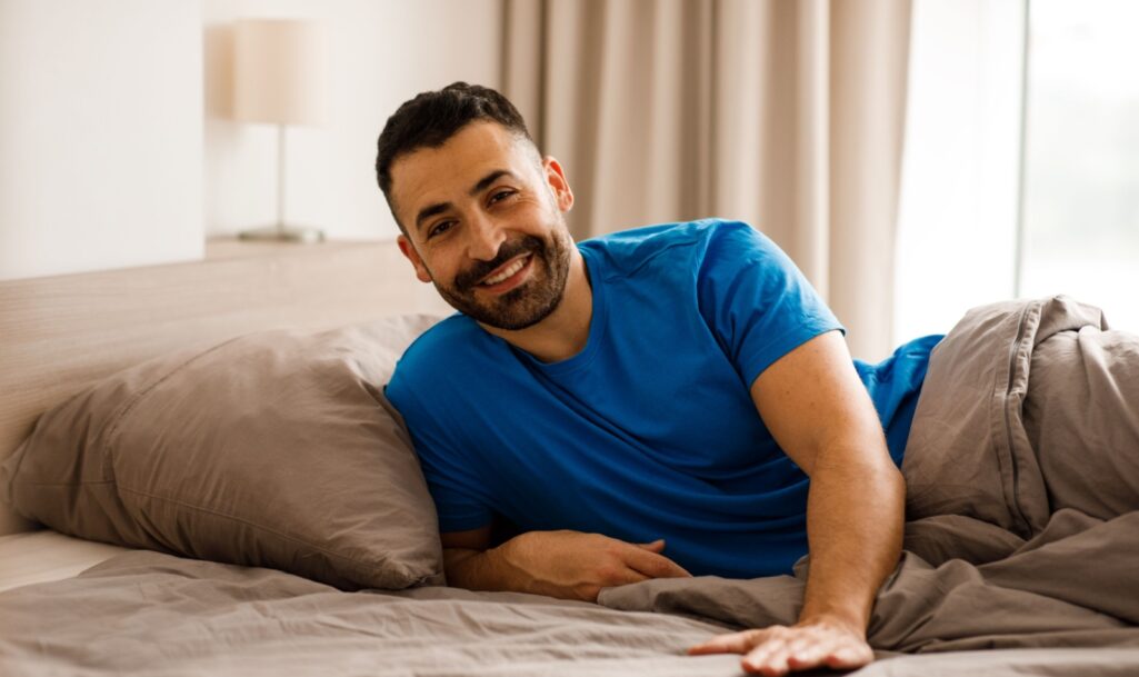 A handsome young man in blue pajamas is lying in bed, touching the empty space next to him and smiling at the camera while waiting for his girlfriend or wife. High testosterone levels consept on a guy.