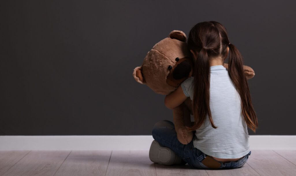 Child abuse. Upset little girl with teddy bear sitting on floor near gray wall indoors, back view.