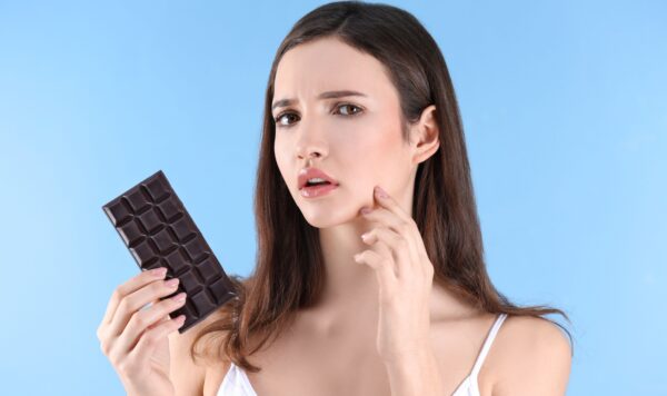 Teenage girl with acne problem holding chocolate bar against color background